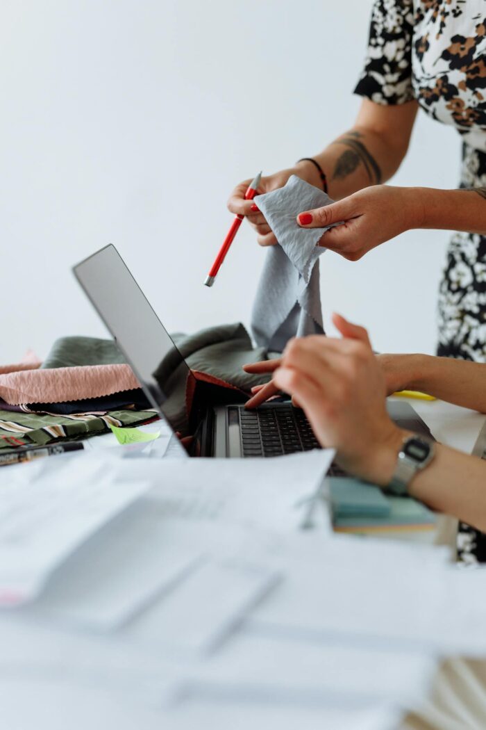 Two female fashion designers collaborating with laptop and fabric samples in a modern studio.